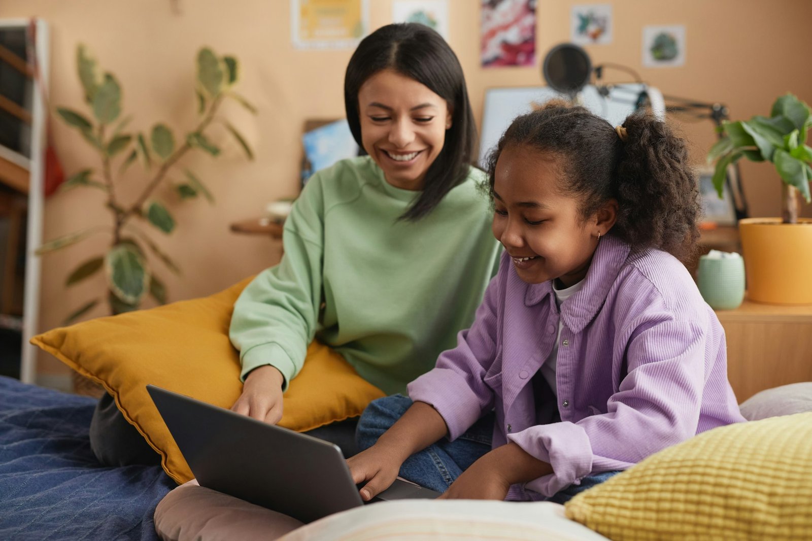 Black mother and daughter using laptop sitting on bed in childs room and smiling